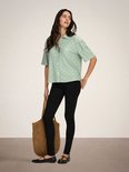 Model in een lichtgroene blouse met broderie anglaise, korte mouwen en knopen, met zwarte skinny jeans en een rieten tas.