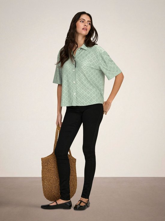 Model in een lichtgroene blouse met broderie anglaise, korte mouwen en knopen, met zwarte skinny jeans en een rieten tas.