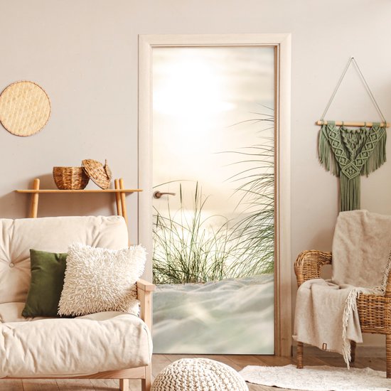 Deurposter met zandduinen en grassprieten bij zonsondergang, in een woonkamer met bank en rieten stoel.