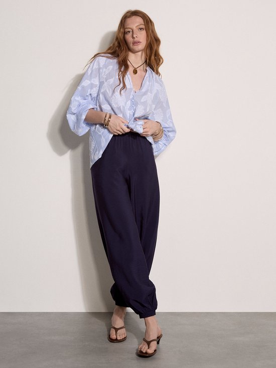 Model in losvallende blauw gestreepte blouse met bloemenpatroon en donkere ballonbroek, staand.