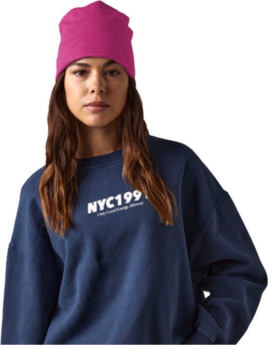Een vrouw met lang bruin haar draagt een marineblauwe trui met opdruk 'NYC199 Only Good Energy Allowed' en een felroze beanie.