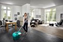 Femme nettoyant le sol avec une serpillière et un seau turquoise dans un salon moderne.