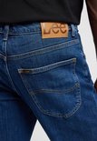 Close-up van de achterkant van een blauwe Lee jeans, met een bruin Lee logo op de tailleband.