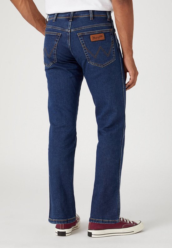 Man in donkerblauwe Wrangler jeans met bruin label, gedragen met bordeauxrode sneakers.