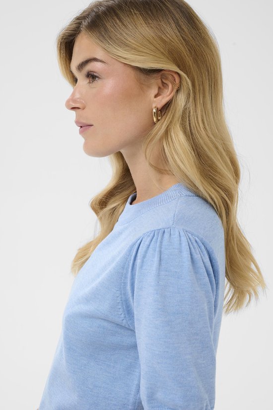 Close-up van een blonde vrouw met blonde golvende haren, gouden oorbel en een lichtblauwe trui.