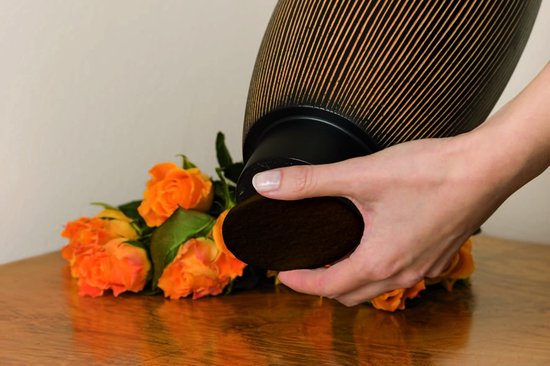 Een hand met een bruine, ronde viltpad die aan de onderkant van een zwarte vaas met gouden ribbels wordt bevestigd, naast oranje rozen.