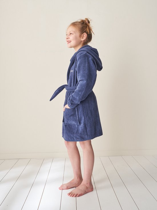 Meisje met het haar in een knot draagt een donkerblauwe badjas met capuchon, ceintuur en geborduurde tekst.