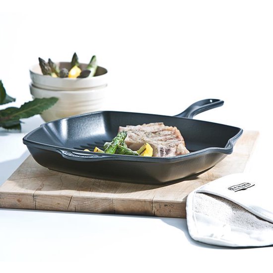 Zwarte gietijzeren grillpan met een stuk vlees en asperges erop, naast een schaal met groenten.