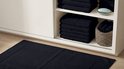 Zwarte badmat op een houten vloer, naast opbergkasten met gestapelde zwarte handdoeken.