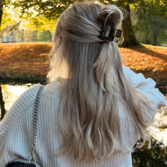 Achterkant van een blonde vrouw met half opgestoken haar in een bruine haarklem, met uitzicht op herfstbomen.