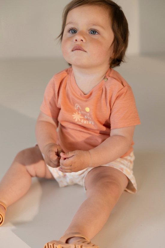 Een jong kind zit en draagt een oranje t-shirt met "LITTLE SUN SHINE" print en een witte broek met gele ruitjes.