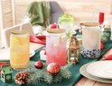 4 glazen met houten deksels en rietjes, gevuld met verschillende gekleurde drankjes, staan op een gedekte tafel met kerstdecoratie.