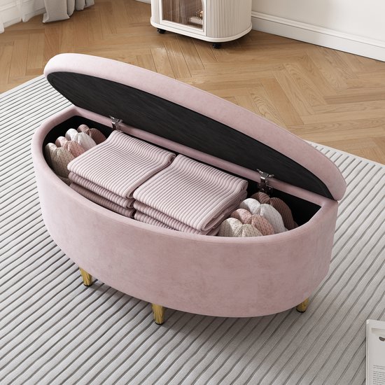 Banc de rangement en velours rose à pieds dorés, le couvercle est ouvert, révélant l'espace de rangement rempli de vêtements.