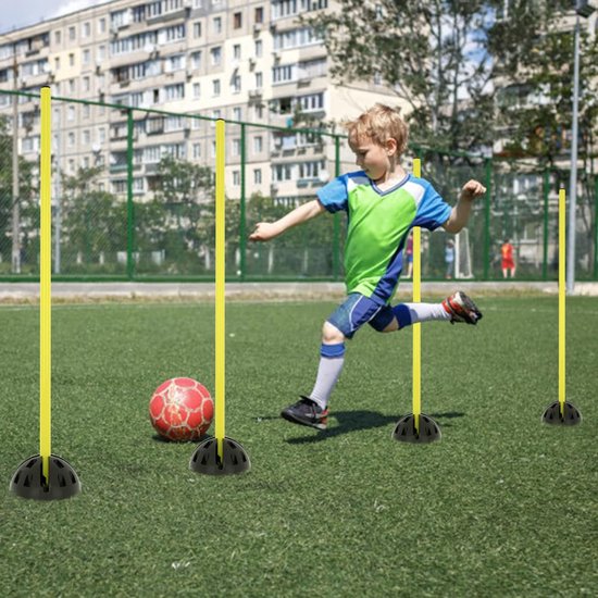 Jonge jongen dribbelt een voetbal langs gele trainingspalen op een grasveld.