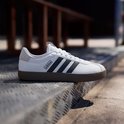 Witte Adidas sneaker met zwarte strepen en bruine zool, op een metalen trap.