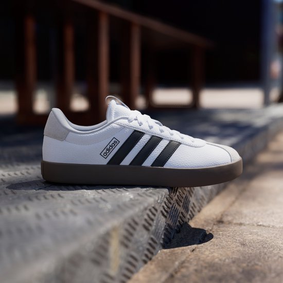 Witte Adidas sneaker met zwarte strepen en bruine zool, op een metalen trap.