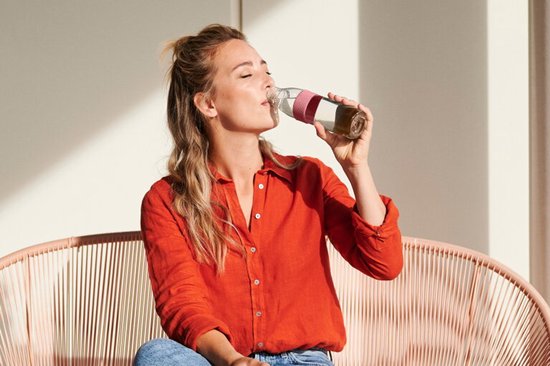 Vrouw met blond haar in een oranje blouse die uit een transparante fles met roze band drinkt, zittend op een roze bank.