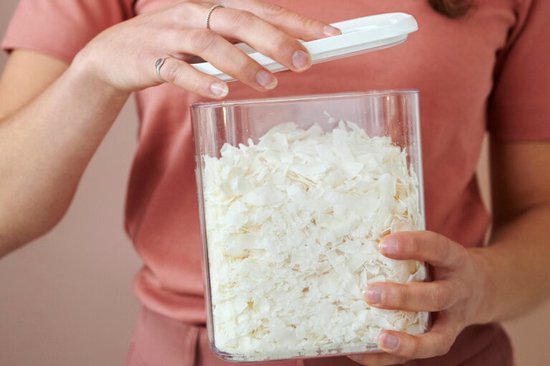 Une femme tient une boîte de conservation transparente avec couvercle blanc, remplie de flocons de noix de coco.