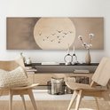 Lange wanddecoratie met een grote, licht beige cirkel tegen een beige achtergrond. Een groepje donkere vogels vliegt in formatie voor de cirkel. Hangt boven een dressoir met boeken en decoraties.
