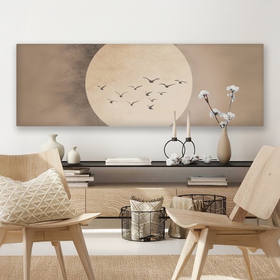Lange wanddecoratie met een grote, licht beige cirkel tegen een beige achtergrond. Een groepje donkere vogels vliegt in formatie voor de cirkel. Hangt boven een dressoir met boeken en decoraties.