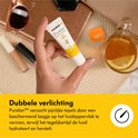 Een hand houdt een kleine tube Medela Purelan Lanolin cream vast, met een kop thee en zonnebril op de achtergrond.