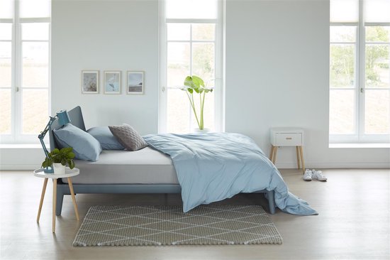 Drap housse jersey gris clair sur un lit avec de la literie bleue, des coussins décoratifs et une table de chevet.