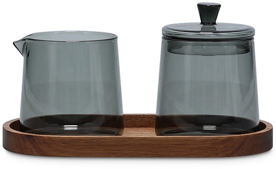 Een grijs glazen melkkannetje en een bijpassende suikerpot met deksel op een ovale houten schaal.