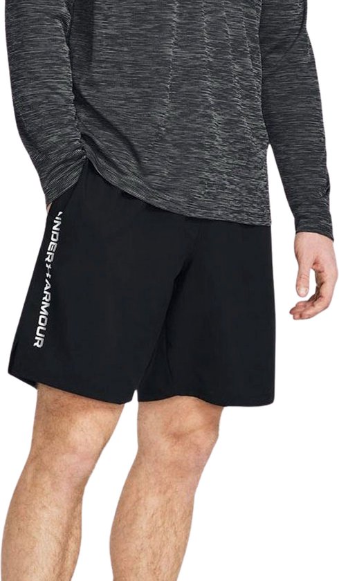 Zwarte Under Armour sportbroek met witte 'UNDER ARMOUR' tekst op de linker broekspijp. Model met de hand in de zak.