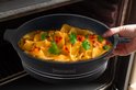 Een hand wordt gebruikt om een zwarte Royalty Line ovenschotel met pasta en kaas uit een oven te halen.