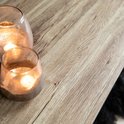 Detailopname van het licht houten tafelblad met een grof patroon. Twee glazen windlichten met brandende kaarsen.