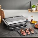 Gesloten Tefal contactgrill met 3 stukken gegrild vlees ernaast. De grill heeft een zilverkleurige afwerking.
