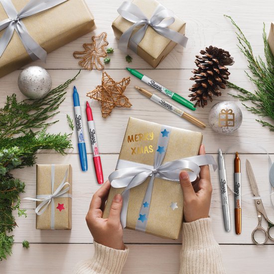 Een hand die een cadeau wikkelt met een gouden Sharpie metallic stift. Een kerstversierde tafel met cadeaus en versieringen.