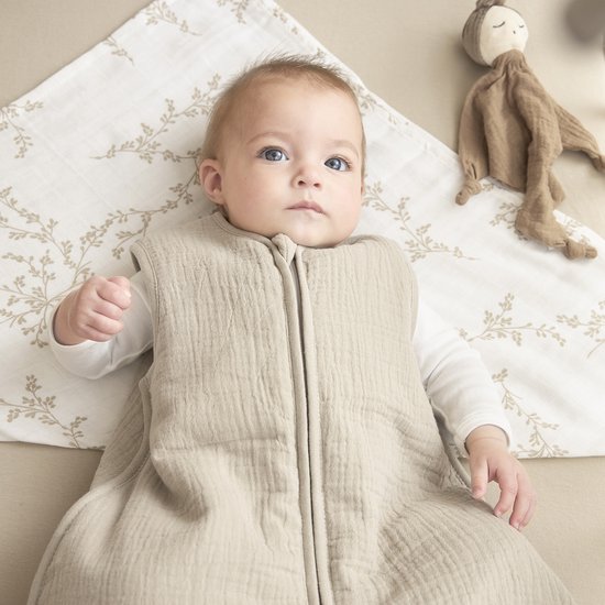 Baby in een zandkleurige, gevoerde slaapzak met rits. Ligt op een laken met takjesprint.