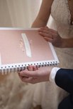 Een man en een bruid houden een open ringband-planner vast. De kaft is koraalkleurig met tekst.