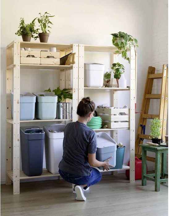 Une femme prend une poubelle grise avec couvercle d'une étagère en bois. Il y a aussi des poubelles bleu clair, vert clair et blanches.