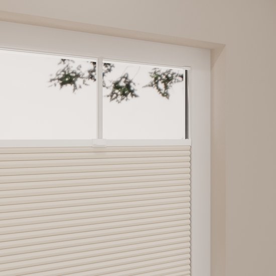 Vue de dessus d'un store plissé couleur sable dans un cadre blanc, avec du feuillage à l'extérieur.