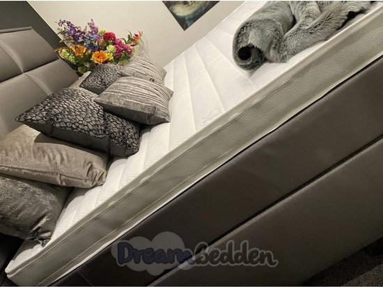 Grijze boxspring met gestoffeerd hoofdbord, witte matras met Aegis-logo, sierkussens en een grijze, pluizige deken.