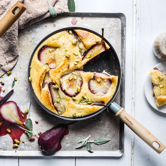 Perencake in een blauwgrijze koekenpan op een metalen bakplaat, bestrooid met pistachenoten. Een stukje cake ligt ernaast.