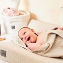 Een lachende baby, gewikkeld in een beige badcape met capuchon. Op de achtergrond staan manden.