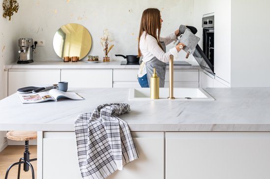 Een vrouw met een schort in een keuken met witte aanrechten en een oven. Ze draagt grijze pannenlappen.