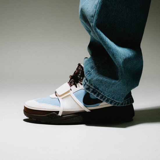 Een persoon draagt een lichtblauwe, beige en bruine sneaker met klittenbandsluiting en jeans.