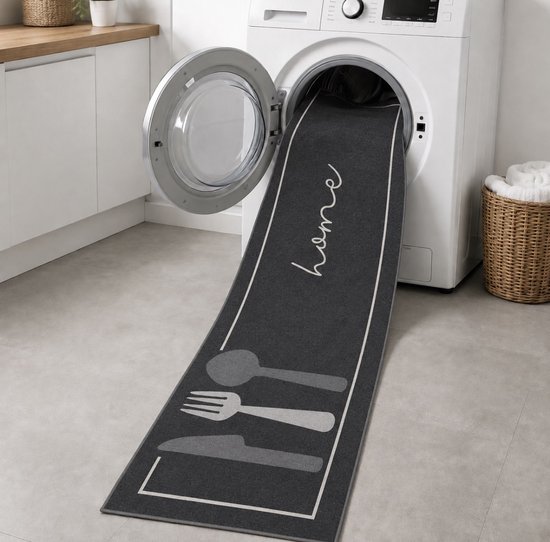 Donkergrijze keukentapijt met 'home' tekst en bestekprint, wordt in wasmachine geplaatst.