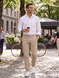 Man loopt over straat met een beige chino broek, witte linnen blouse, witte sneakers en een koffiebeker.