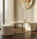 Stijlvolle woonkamer met ovale salontafel met marmerlook blad en gouden frame, twee crèmekleurige poefs, en twee mokken.