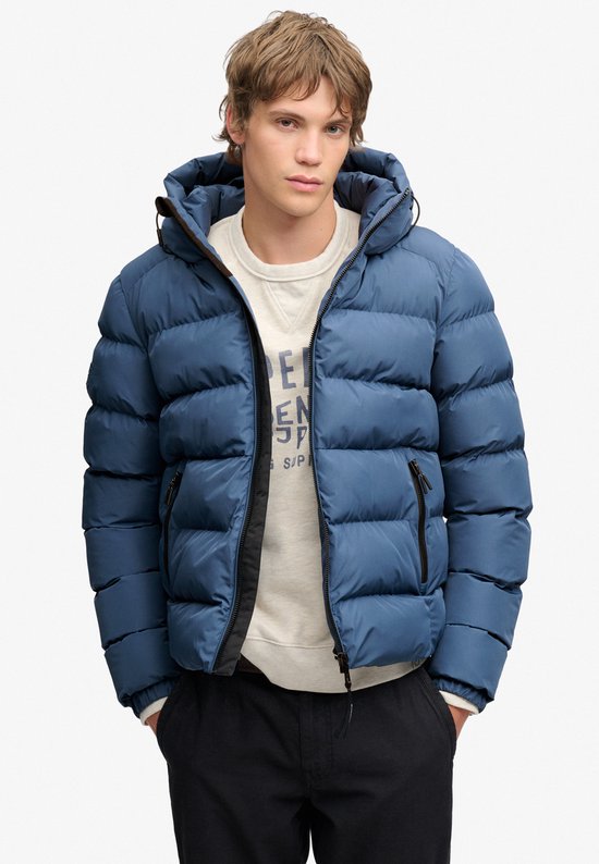 Man met blauwe, gewatteerde Superdry jas met capuchon, wit T-shirt en zwarte broek.