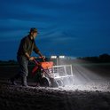 Een man met een hoed bewerkt een akker met een motorisch aangedreven ploeg, verlicht door twee LED-lampen op de machine.