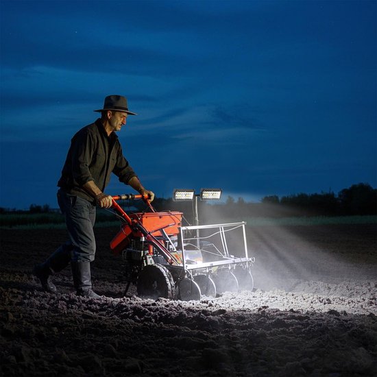 Een man met een hoed bewerkt een akker met een motorisch aangedreven ploeg, verlicht door twee LED-lampen op de machine.