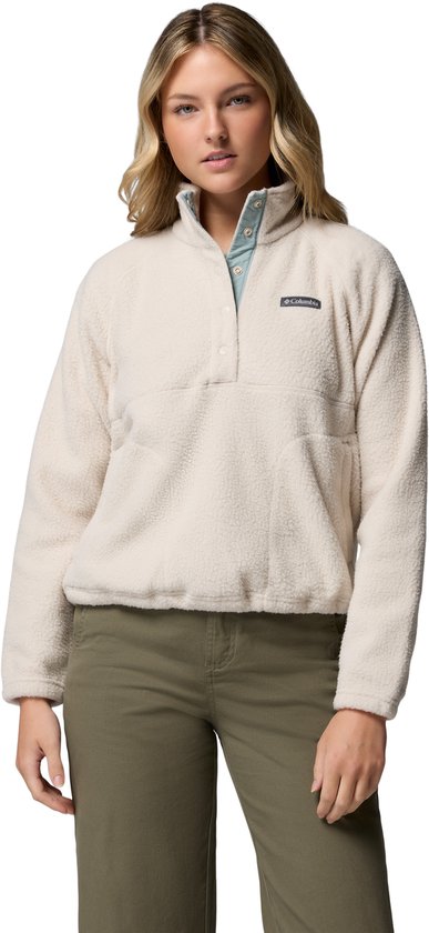 Een vrouw draagt een gebroken witte, teddyfleece trui met hoge hals, drukknoopsluiting en lichtblauwe kraag. Ze draagt een olijfgroene broek.