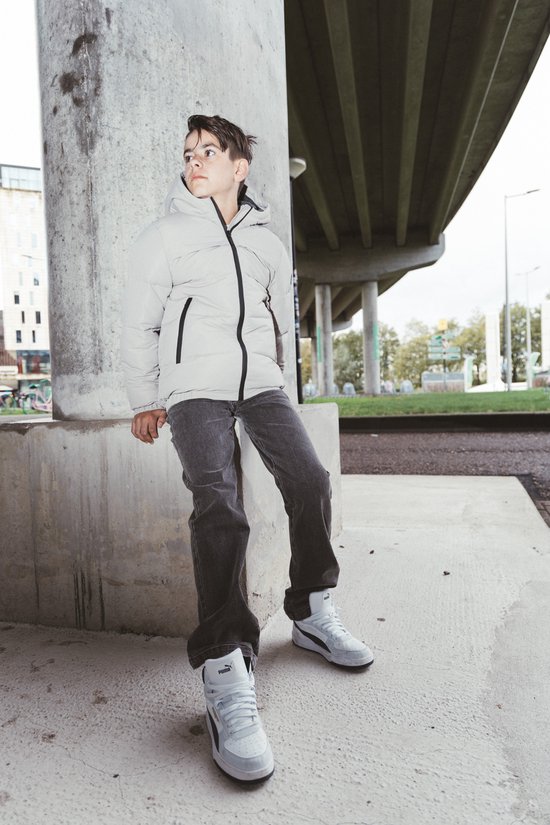 Jongen in een lichtgrijze winterjas met capuchon, zittend tegen een betonnen pilaar, kijkend naar rechts.