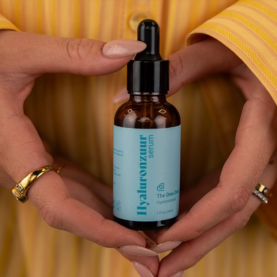 Handen met lichte nagellak houden glazen flesje Hyaluronzuur serum vast. Gouden ringen aan de vingers.